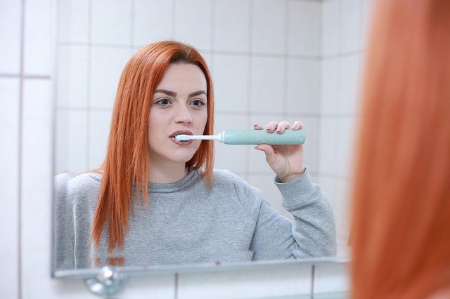 Redheaded woman using an electric toothbrush in the mirror.