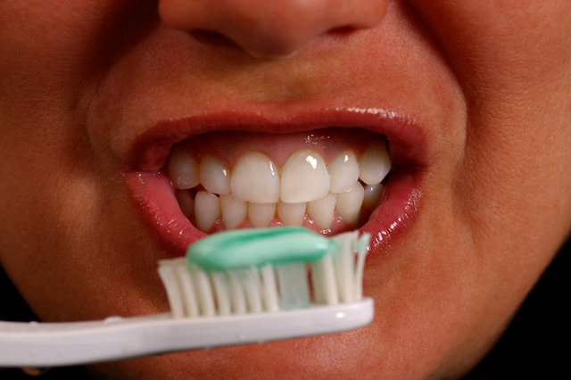 A close up photo of a person smiling, holding a toothbrush with toothpaste on it in front of their mouth.